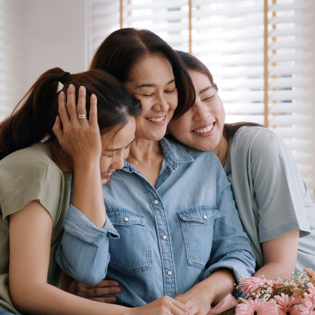 Seorang ibu dan dua anak perempuannya saling berpelukan dengan penuh kehangatan, melambangkan keharmonisan dan pemahaman yang tumbuh melalui Tes Mind Modes™ Keluarga.